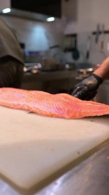 Şef, profesyonel bir mutfakta taze somon fileto dilimliyor. Siyah eldivenli ve çiğ balık desenli eller..