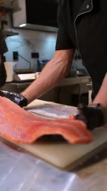 Şef, profesyonel bir mutfakta taze somon fileto dilimliyor. Siyah eldivenli ve çiğ balık desenli eller..