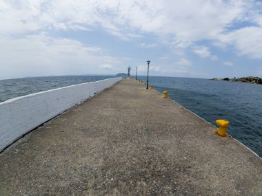 Parçalı bulutlu bir gökyüzünün altında durgun mavi denize uzanan sarı demirli direkler ve sokak lambalarıyla uzun beton iskele..
