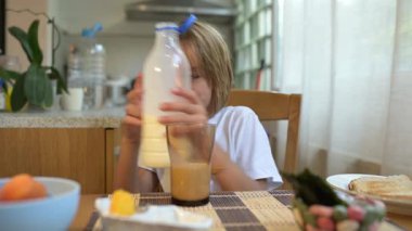 Küçük çocuk sıcak mutfakta kahvaltı masasında süt içiyor. Doğal ışık ve sağlıklı yiyeceklerle huzurlu bir sabah rutini..