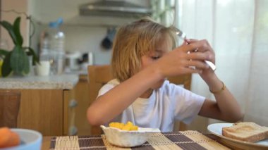 Genç bir çocuk kahvaltı masasında çatalla taze meyve yiyor. Doğal ışık ve sağlıklı yiyeceklerle huzurlu bir sabah ortamı..