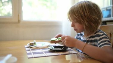Küçük bir çocuk kahvaltı masasında sade ekmek yiyor. Doğal ışık ve basit ev yapımı yiyeceklerle huzurlu bir sabah rutini..
