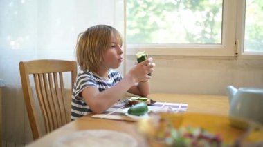 Evde yemek masasında salatalık ve ekmek yiyen küçük bir çocuk. Doğal gün ışığı, sakin atmosfer, günlük aile ortamında sağlıklı ve basit atıştırmalıklar.