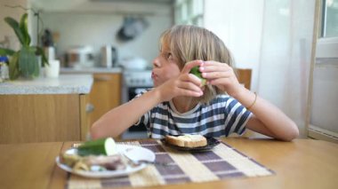 Evde yemek masasında salatalık ve ekmek yiyen küçük bir çocuk. Doğal gün ışığı, sakin atmosfer, günlük aile ortamında sağlıklı ve basit atıştırmalıklar.