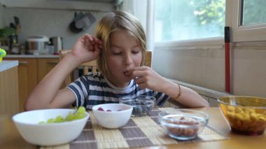 Evde yemek masasındaki bir kaseden yabanmersini toplayan küçük bir çocuk. Doğal gün ışığı, rahat bir atmosfer, sıcacık mutfakta meyveli ve fındıklı sağlıklı atıştırmalıklar..