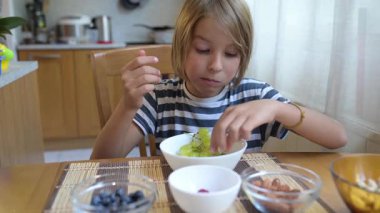 Evde yemek masasında yeşil üzüm yiyen küçük bir çocuk. Doğal ışık, gevşek mutfak seti, fındık ve böğürtlenli sağlıklı meyve aperatifi.