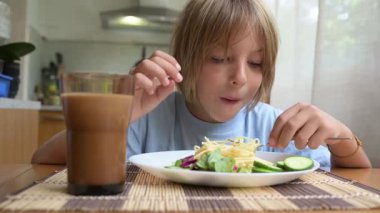 Küçük bir çocuk mutfak masasında taze sebzelerle spagetti yiyor. Sıcak atmosfer, doğal ışık, sağlıklı öğle yemeği ve her gün içilen içki..