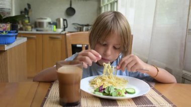 Küçük bir çocuk mutfak masasında taze sebzelerle spagetti yiyor. Sıcak atmosfer, doğal ışık, sağlıklı öğle yemeği ve her gün içilen içki..