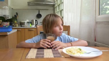 Küçük çocuk sıcak mutfakta kahvaltı masasında süt içiyor. Doğal ışık ve sağlıklı yiyeceklerle huzurlu bir sabah rutini..