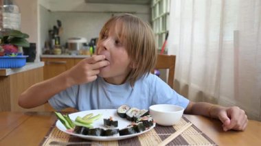 Evde mutfak masasında suşi yiyen küçük bir çocuk. Sıcak atmosfer, doğal ışık, geleneksel Asya yemekleri, sağlıklı öğle yemeği veya günlük aile ortamında akşam yemeği.