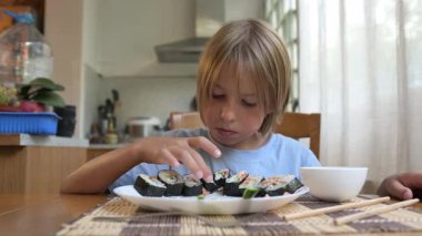 Evde mutfak masasında suşi yiyen küçük bir çocuk. Sıcak atmosfer, doğal ışık, geleneksel Asya yemekleri, sağlıklı öğle yemeği veya günlük aile ortamında akşam yemeği.