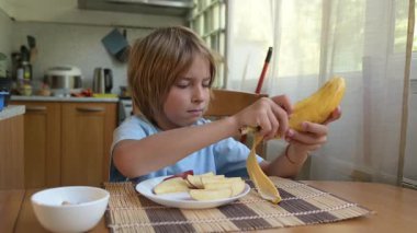 Evde mutfak masasında muz ve elma dilimleri yiyen küçük bir çocuk. Doğal ışık, sağlıklı meyve atıştırmalığı, sakin ve rahat günlük atmosfer.