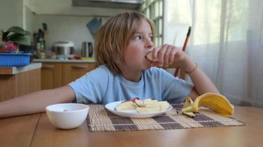 Evde mutfak masasında muz ve elma dilimleri yiyen küçük bir çocuk. Doğal ışık, sağlıklı meyve atıştırmalığı, sakin ve rahat günlük atmosfer.
