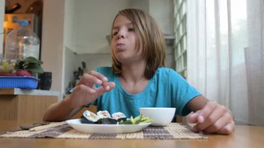 Evde mutfak masasında suşi yiyen küçük bir çocuk. Sıcak atmosfer, doğal ışık, geleneksel Asya yemekleri, sağlıklı öğle yemeği veya günlük aile ortamında akşam yemeği.