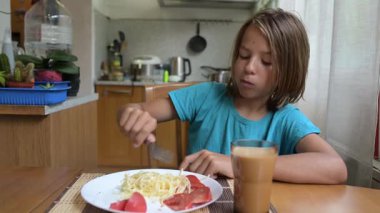 Küçük bir çocuk mutfak masasında taze sebzelerle spagetti yiyor. Sıcak atmosfer, doğal ışık, sağlıklı öğle yemeği ve her gün içilen içki..