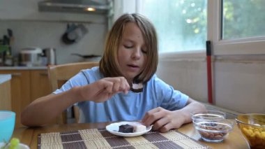 Evde mutfak masasında kremalı tatlı yiyen küçük bir çocuk. Rahat bir atmosfer, doğal ışık, rahat ortamlarda tatlı süt ürünleri ikram edilen günlük atıştırma zamanı..