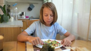 Küçük bir çocuk mutfak masasında taze sebze salatası yiyor. Doğal gün ışığı, sağlıklı yaşam tarzı, rahat günlük atmosferde basit ev yapımı yemek..