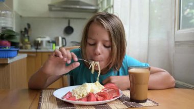 Küçük bir çocuk mutfak masasında taze sebzelerle spagetti yiyor. Sıcak atmosfer, doğal ışık, sağlıklı öğle yemeği ve her gün içilen içki..