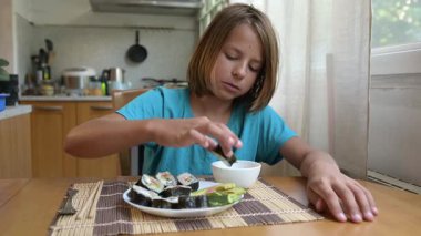 Evde mutfak masasında suşi yiyen küçük bir çocuk. Sıcak atmosfer, doğal ışık, geleneksel Asya yemekleri, sağlıklı öğle yemeği veya günlük aile ortamında akşam yemeği.