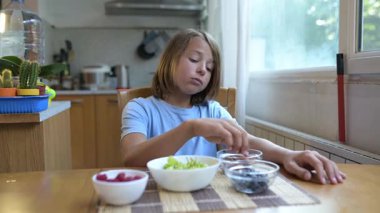 Mutfak masasında badem yiyen küçük bir çocuk. Doğal gün ışığı, sağlıklı çerez, basit ve rahat günlük atmosfer, masada meyve ve kaseler..