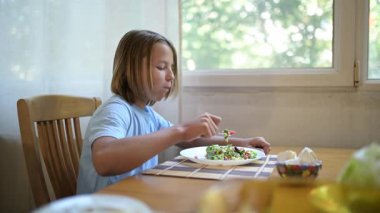 Küçük bir çocuk mutfak masasında taze sebze salatası yiyor. Doğal gün ışığı, sağlıklı yaşam tarzı, rahat günlük atmosferde basit ev yapımı yemek..