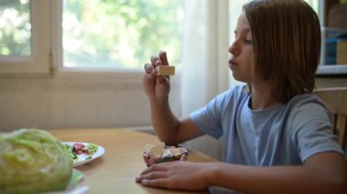 Evde mutfak masasında peynir yiyen küçük bir çocuk. Doğal gün ışığı, salata ve sebzeli sağlıklı aperatif, rahat ve basit günlük atmosfer..