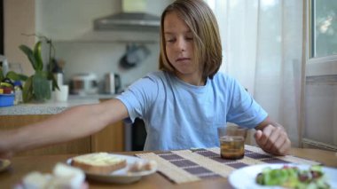 Küçük bir çocuk mutfak masasında bardağa limonata dolduruyor. Doğal gün ışığı, ferahlatıcı içecek, basit günlük yemeklerle sıcak aile atmosferi..