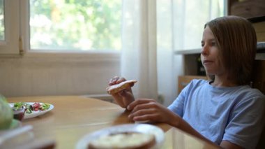 Küçük bir çocuk mutfak masasında sandviç yiyor. Doğal gün ışığı, basit ev yapımı aperatif yanında salata, rahat ve günlük atmosfer..