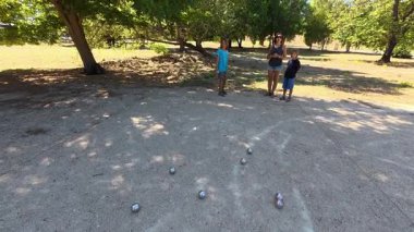 Aile parkta birlikte petanque oynuyor. Yetişkinler ve çocuklar güneşli bir yaz gününde sırayla metal toplar atıyorlar. Eğlenceli bir açık hava aktivitesi ve aile bağları oyunu.