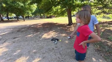 Güneşli bir günde iki çocuk açık havada Petanque oynuyor. Çocuklar sırayla parktaki çakıl taşlarına metal toplar atıyor, eğlence ve spor aktivitelerinin keyfini çıkarıyorlar..