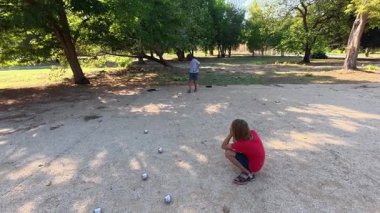 Güneşli bir günde iki çocuk açık havada Petanque oynuyor. Çocuklar sırayla parktaki çakıl taşlarına metal toplar atıyor, eğlence ve spor aktivitelerinin keyfini çıkarıyorlar..