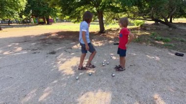 Güneşli bir günde iki çocuk açık havada Petanque oynuyor. Çocuklar sırayla parktaki çakıl taşlarına metal toplar atıyor, eğlence ve spor aktivitelerinin keyfini çıkarıyorlar..