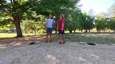 Güneşli bir günde iki çocuk açık havada Petanque oynuyor. Çocuklar sırayla parktaki çakıl taşlarına metal toplar atıyor, eğlence ve spor aktivitelerinin keyfini çıkarıyorlar..