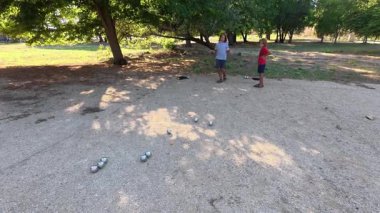 Güneşli bir günde iki çocuk açık havada Petanque oynuyor. Çocuklar sırayla parktaki çakıl taşlarına metal toplar atıyor, eğlence ve spor aktivitelerinin keyfini çıkarıyorlar..