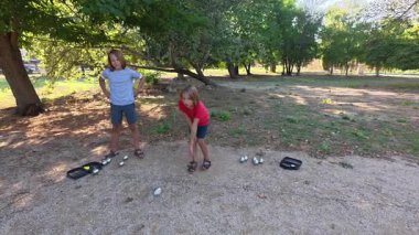 Güneşli bir günde iki çocuk açık havada Petanque oynuyor. Çocuklar sırayla parktaki çakıl taşlarına metal toplar atıyor, eğlence ve spor aktivitelerinin keyfini çıkarıyorlar..
