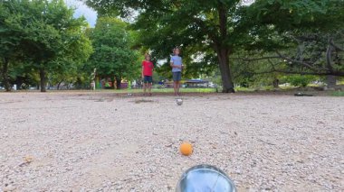 Güneşli bir günde iki çocuk açık havada Petanque oynuyor. Çocuklar sırayla parktaki çakıl taşlarına metal toplar atıyor, eğlence ve spor aktivitelerinin keyfini çıkarıyorlar..
