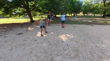 Aile parkta birlikte petanque oynuyor. Yetişkinler ve çocuklar güneşli bir yaz gününde sırayla metal toplar atıyorlar. Eğlenceli bir açık hava aktivitesi ve aile bağları oyunu.