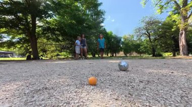 Aile parkta birlikte petanque oynuyor. Yetişkinler ve çocuklar güneşli bir yaz gününde sırayla metal toplar atıyorlar. Eğlenceli bir açık hava aktivitesi ve aile bağları oyunu.
