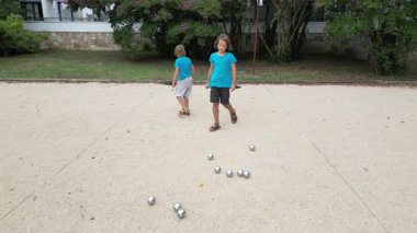 Güneşli bir günde iki çocuk açık havada Petanque oynuyor. Çocuklar sırayla parktaki çakıl taşlarına metal toplar atıyor, eğlence ve spor aktivitelerinin keyfini çıkarıyorlar..