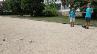 Güneşli bir günde iki çocuk açık havada Petanque oynuyor. Çocuklar sırayla parktaki çakıl taşlarına metal toplar atıyor, eğlence ve spor aktivitelerinin keyfini çıkarıyorlar..