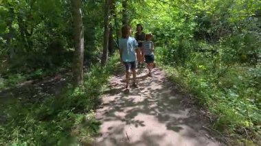 Yeşil bir ormanda aile yürüyüşü. Kadın ve iki çocuk gölgeli bir patika boyunca yürürler, açık hava macerası, doğa keşfi, yaz tatili ve ormanda birlikte zaman geçirmenin tadını çıkarırlar..