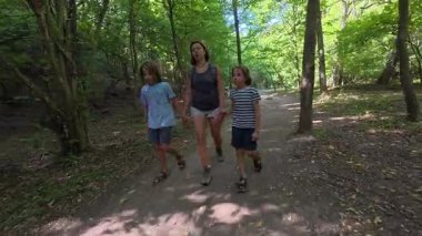Yeşil bir ormanda aile yürüyüşü. Kadın ve iki çocuk gölgeli bir patika boyunca yürürler, açık hava macerası, doğa keşfi, yaz tatili ve ormanda birlikte zaman geçirmenin tadını çıkarırlar..