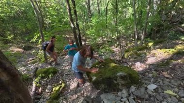 Aile ormanda kayalık bir patikaya tırmanıyor. Çocuklar ve kadınlar birlikte yokuş yukarı yürür, destek için elleri kullanır, açık hava macerasından, takım çalışmasından ve yazın doğayı keşfetmekten zevk alır..