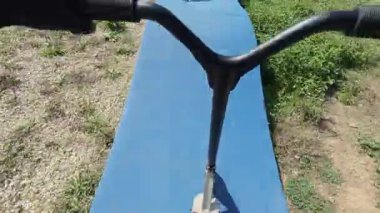 POV shot of riding a red scooter on a blue pump track, hands on handlebars in gloves, active outdoor lifestyle in summer.