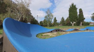 Two children riding scooters on a blue pump track in a park, wearing helmets and protective gear, active outdoor lifestyle on a sunny day.