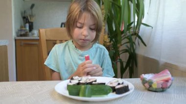 Young boy sitting at the kitchen table holding a crab stick with a plate of sushi rolls and cucumber in front of him.