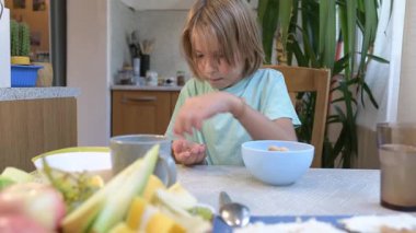 Young boy sitting at the kitchen table peeling and eating peanuts and pistachios with a bowl in front of him.