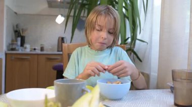 Young boy sitting at the kitchen table peeling and eating peanuts and pistachios with a bowl in front of him.