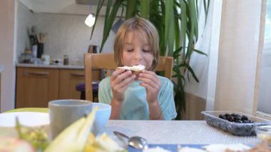 Young boy sitting at the kitchen table eating a sandwich with cream cheese, with grapes and dishes on the table.