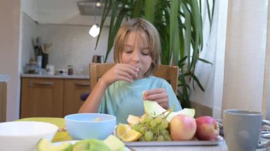 Young boy sitting at the kitchen table eating fresh fruits including orange, grapes, melon, and apples.